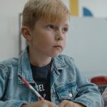 A young boy in a denim jacket attentively learning in a classroom setting.